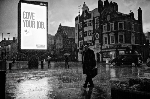Love Your Job. A heavy downpour of rain soaks pedestrians and a businessman, as they pass an illuminated advertising sign saying "Love Your Job". Hammersmith, London. January 14, 2011. Photo: ©Edmond Terakopian